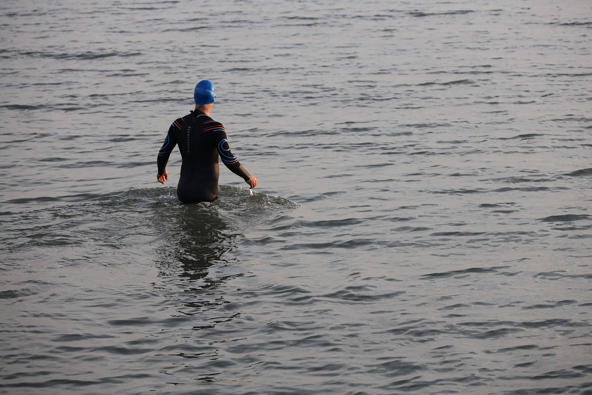 A swimmer in a wetsuit and cap wading out into a grey sea, seen from behind