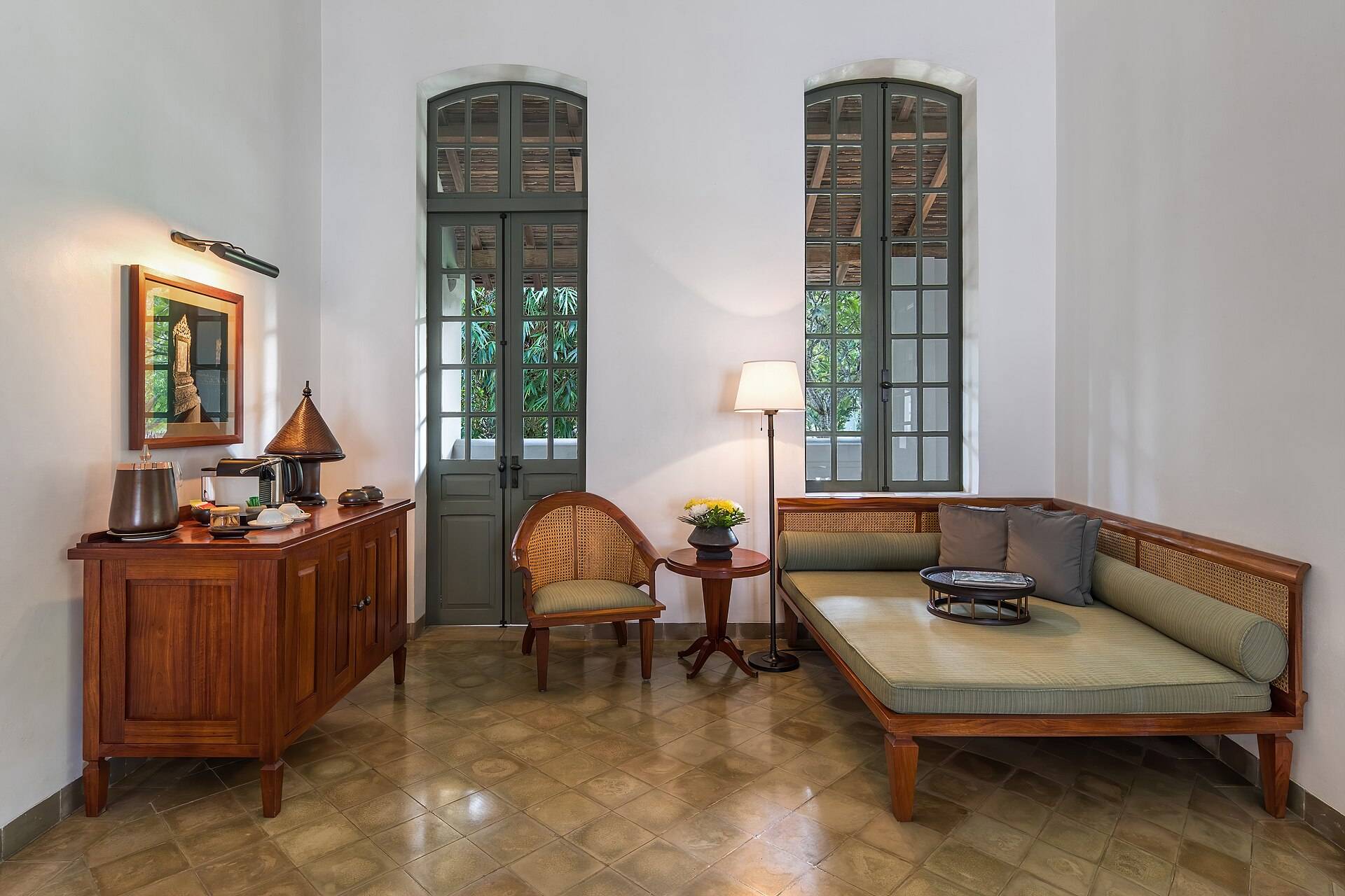A quiet room with a teak daybed, a cane armchair and tall shuttered doors
