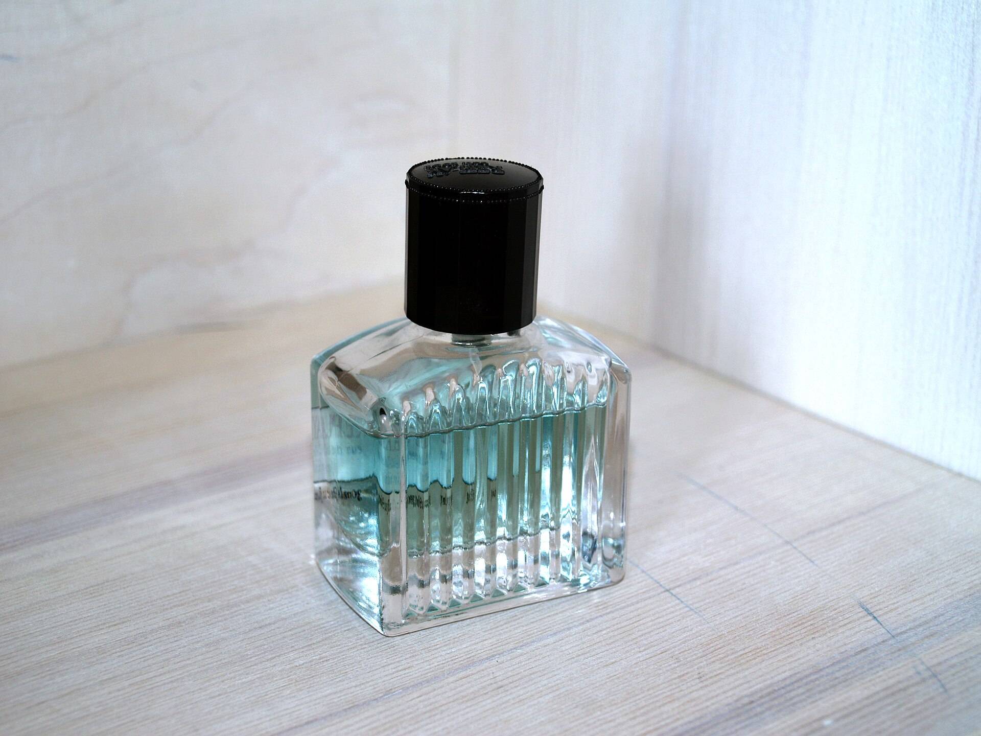 A ribbed glass perfume bottle with a black cap on a pale wooden shelf against a limewashed wall
