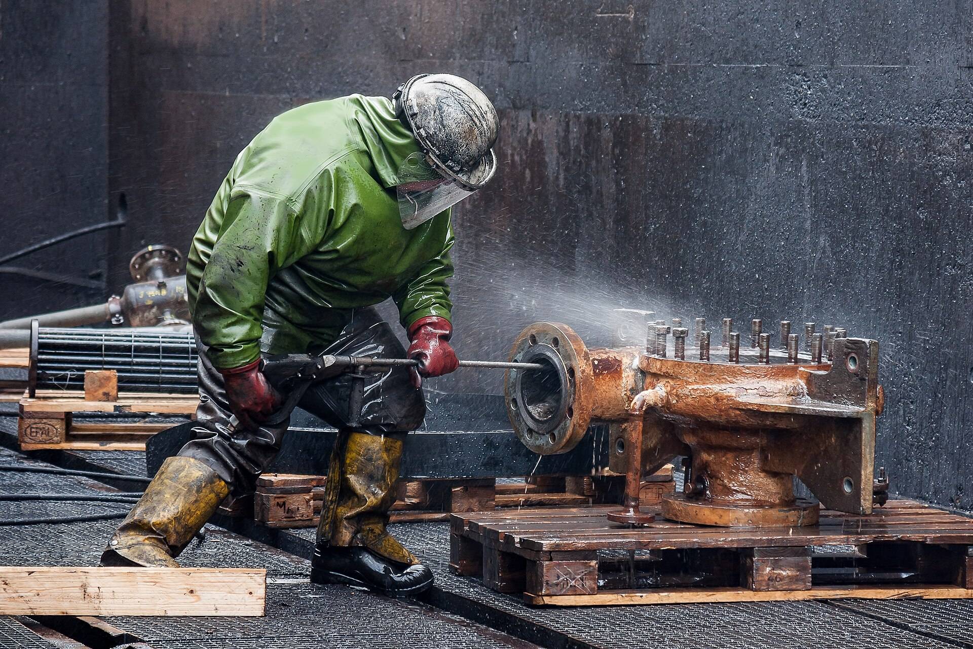 A man in waterproofs pressure washing a rusty machine part on a pallet