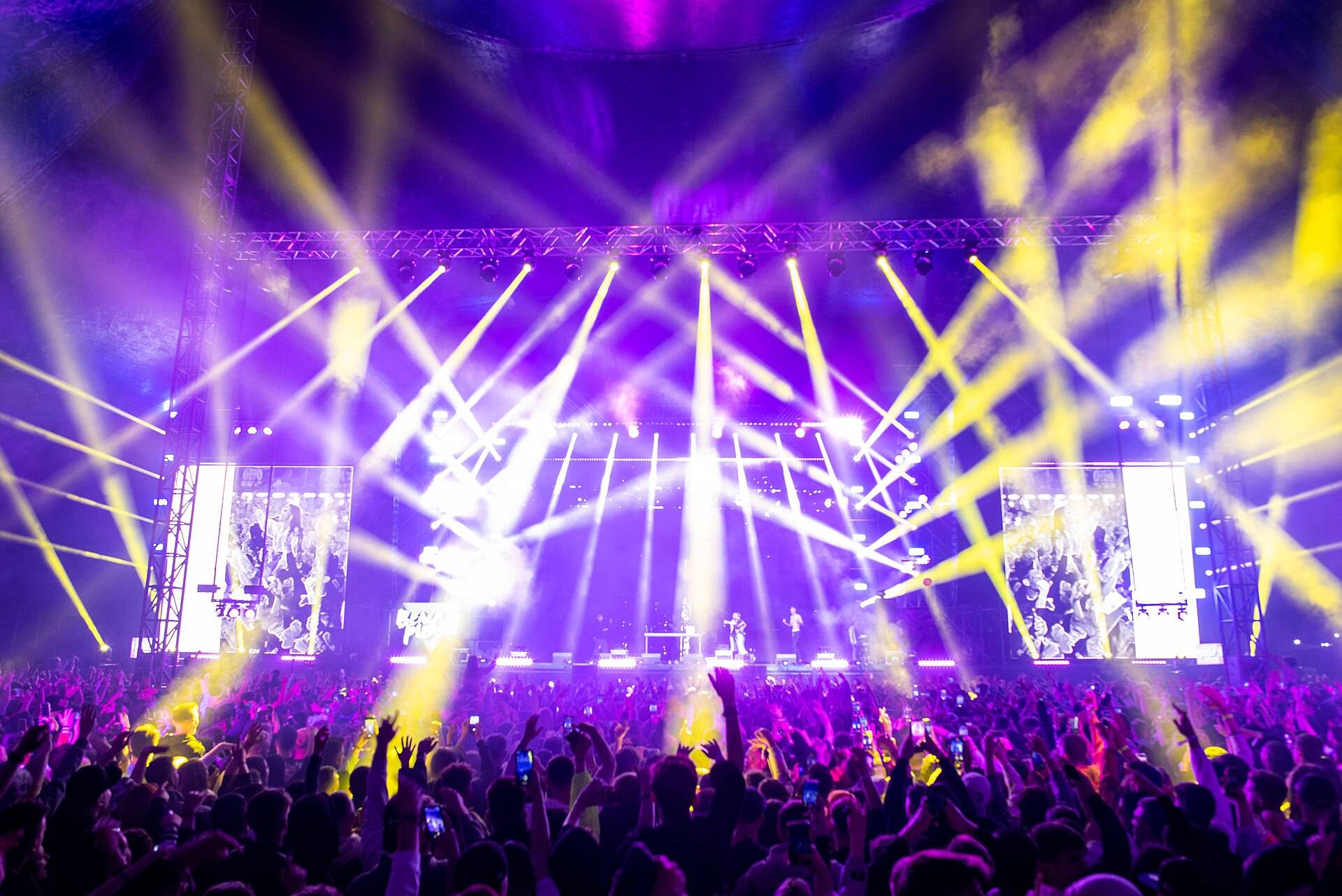 A festival crowd at night facing a stage, light beams fanning out over it between two big screens