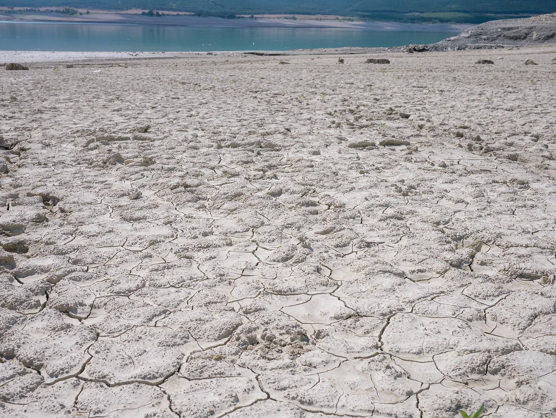 A dried reservoir bed of cracked pale mud running out to a thin line of water