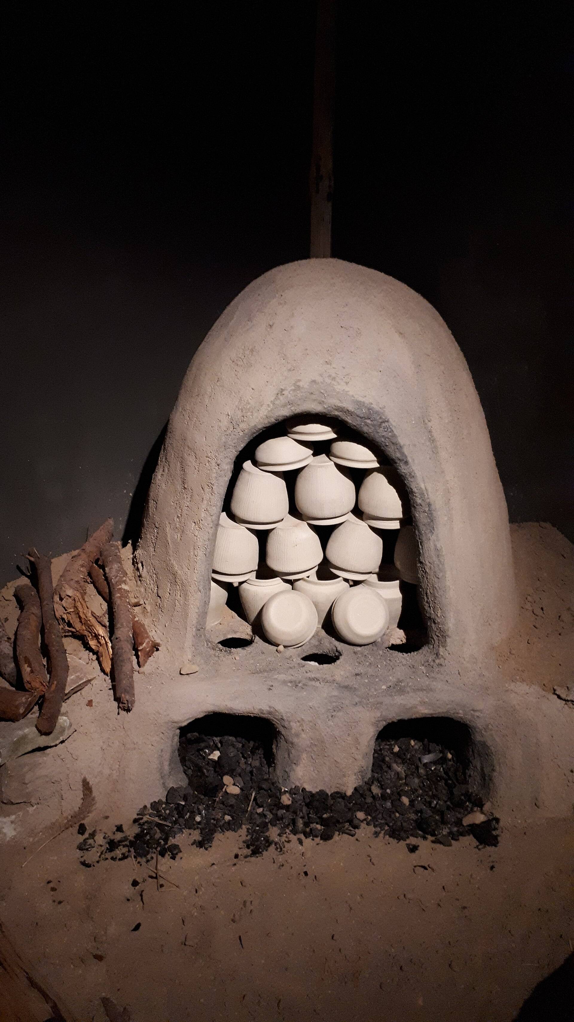 A domed clay kiln stacked with pale pots, lit in a dark room