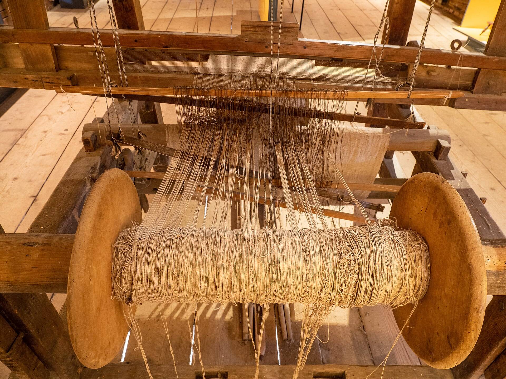 The warp beam of a wooden loom, wound with linen threads running up to the reed