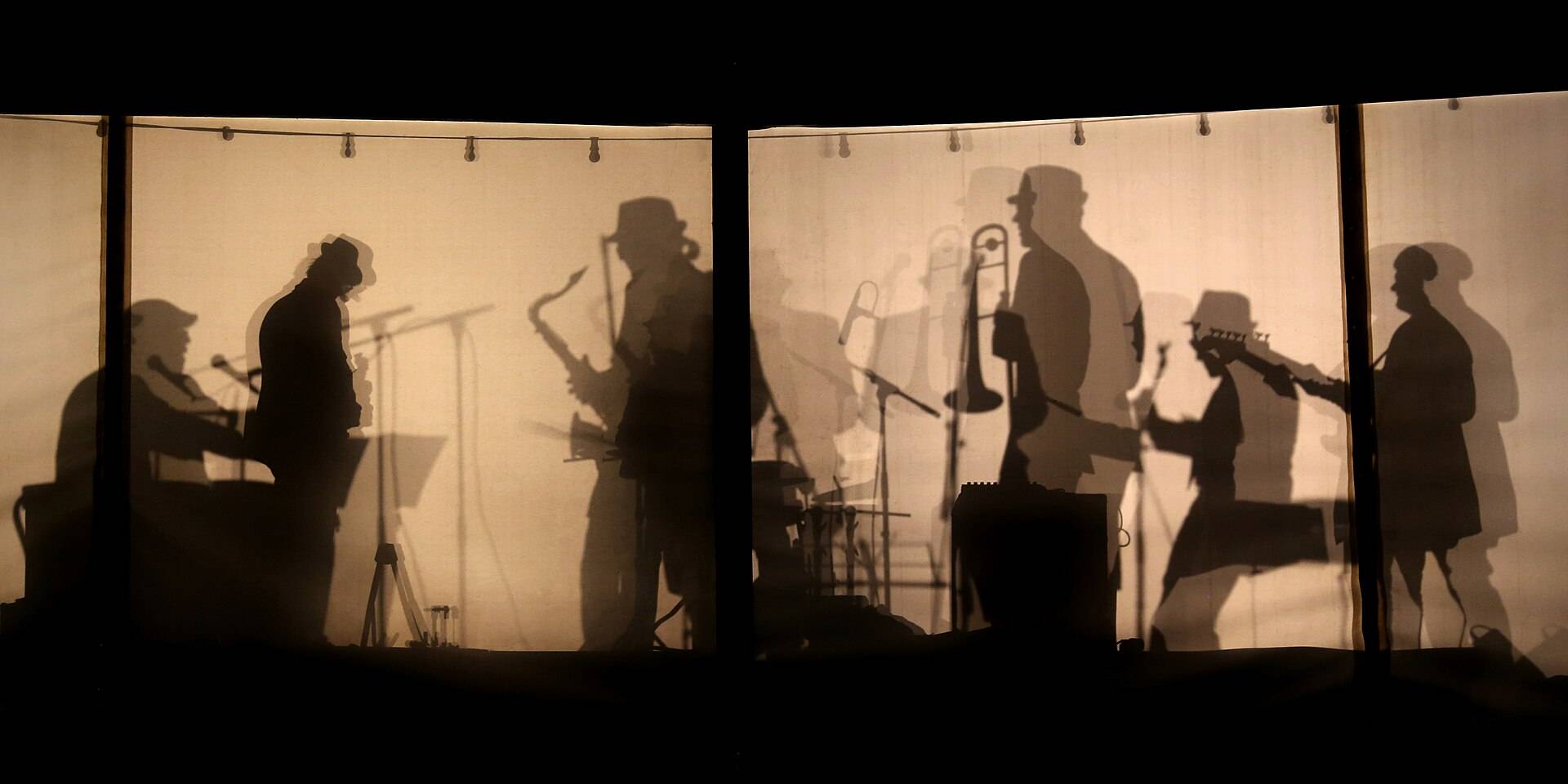 The shadows of a band, horns and all, thrown on a lit screen behind the stage
