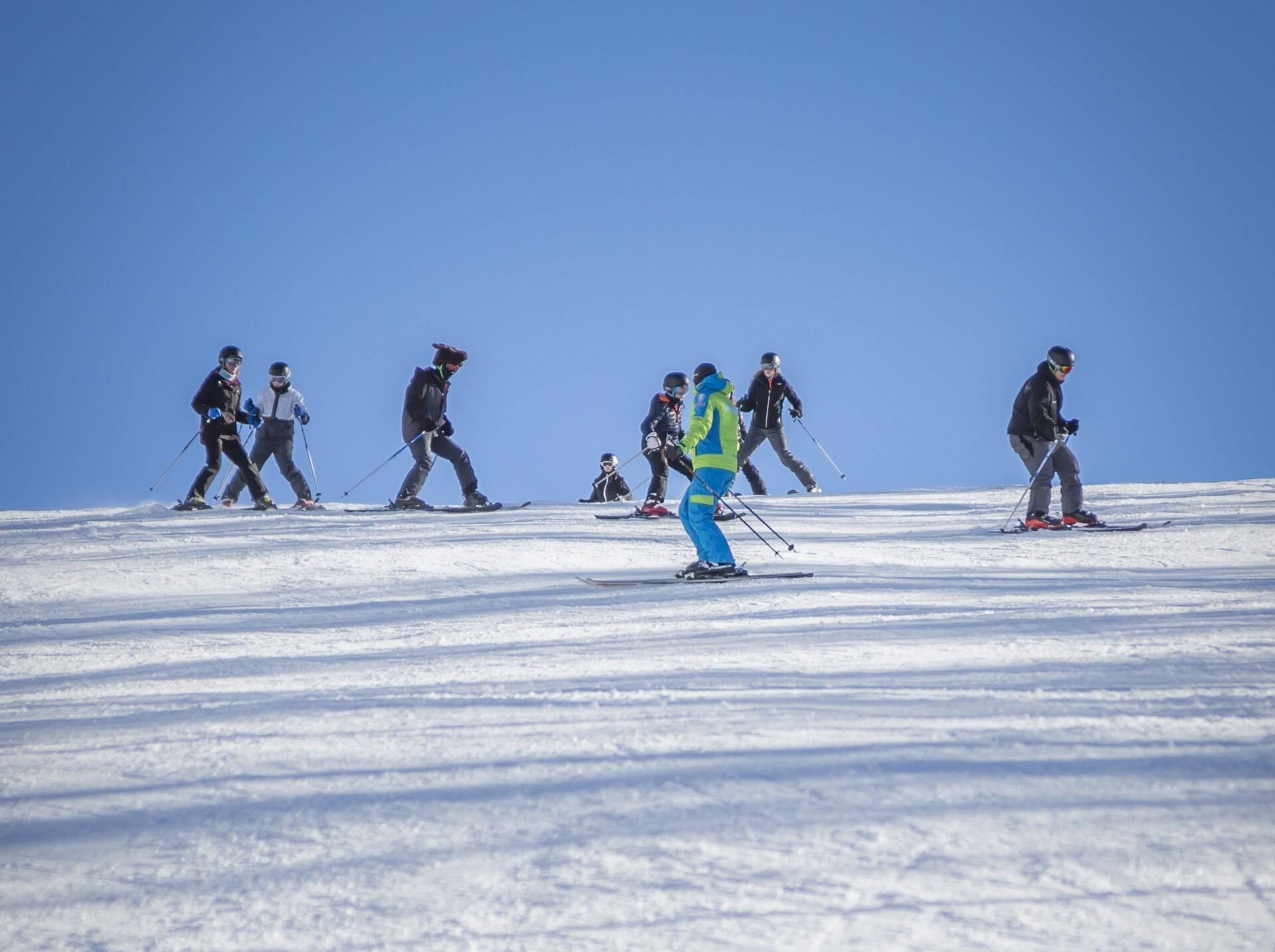 A ski class in a line behind its instructor, crossing a sunlit slope under a blue sky