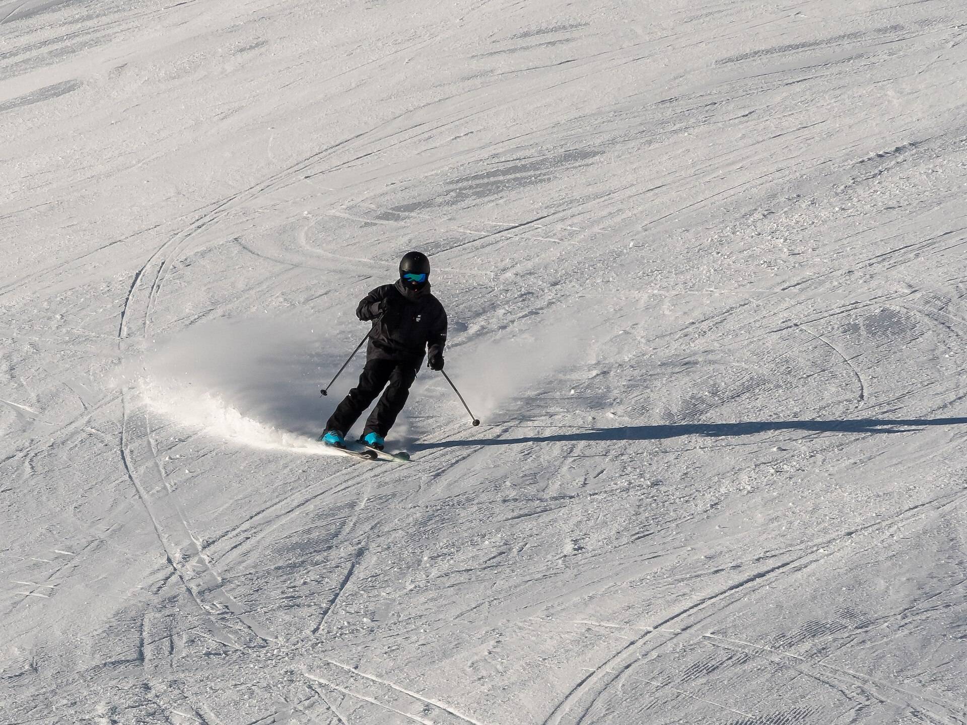 A skier in black carving a turn across a wide groomed slope, spray behind, seen from above