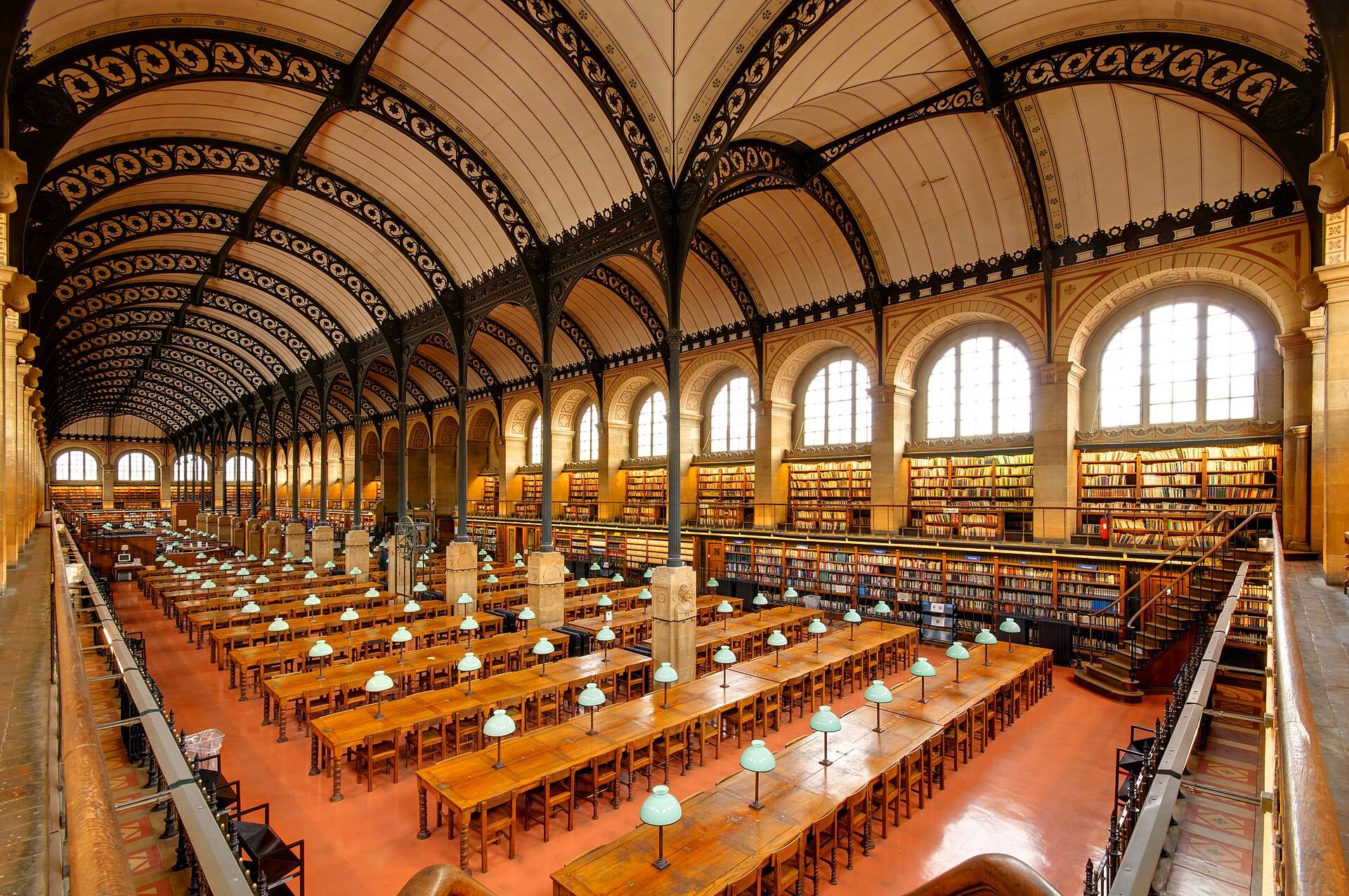 A long vaulted reading room with rows of wooden desks, green lamps and walls of books