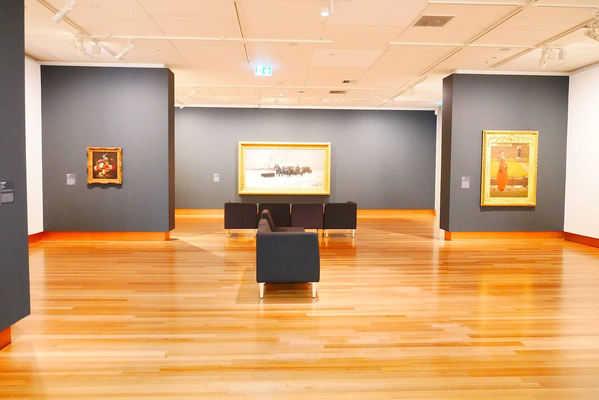 A gallery room with grey walls, framed paintings and a bench on a timber floor