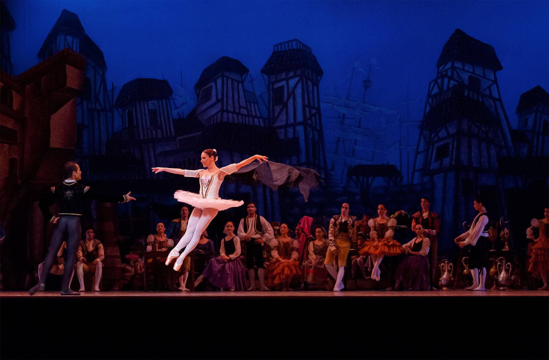 A ballet dancer mid leap on a lit stage, the company and a painted set behind