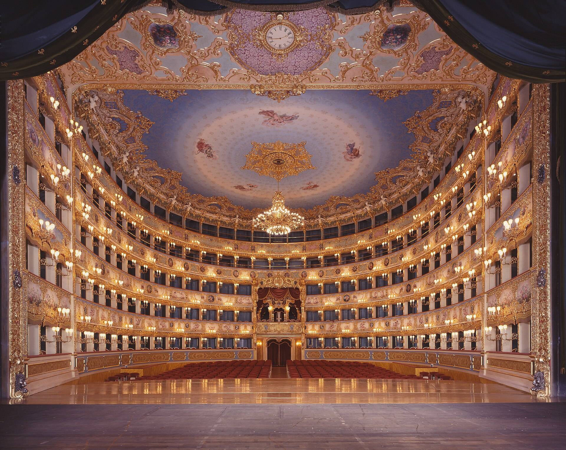 A gilded opera house seen from the stage, tiers of boxes around red seats