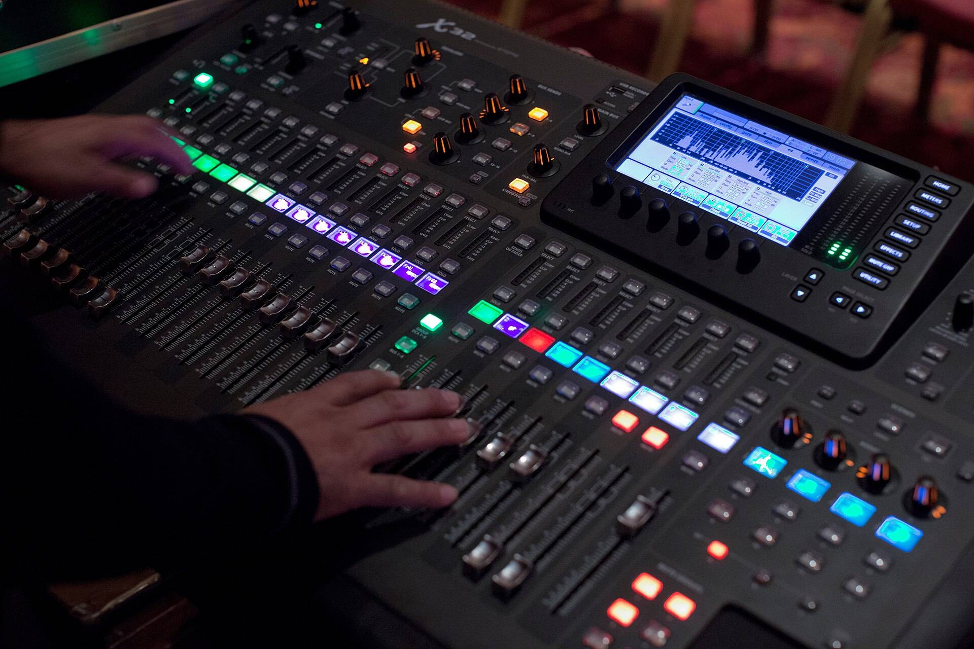 Two hands on the faders of a lit digital mixing desk in a dark booth